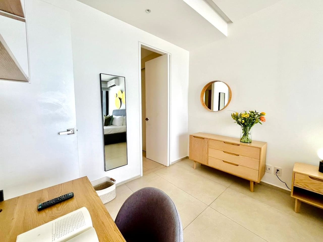 Une chambre moderne avec un bureau en bois, une chaise, une table de chevet, une commode et un miroir au mur.