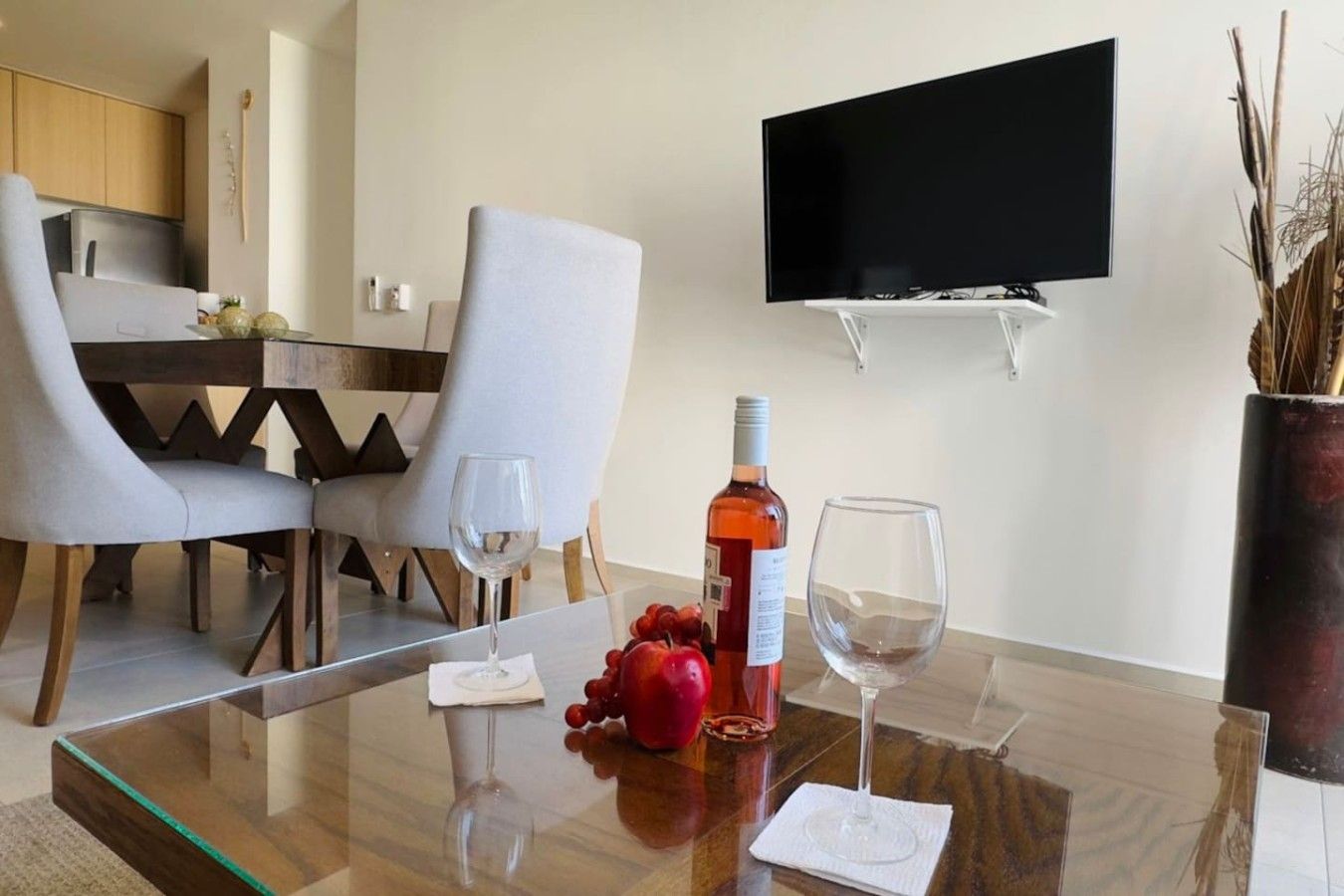 A modern dining area with a glass table, two chairs, a bottle of wine, and a TV mounted on the wall.
