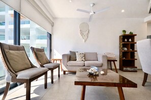 Ein modernes Wohnzimmer mit einer Couch, zwei Sesseln, einem Couchtisch, einem Bücherregal und einem Deckenventilator.