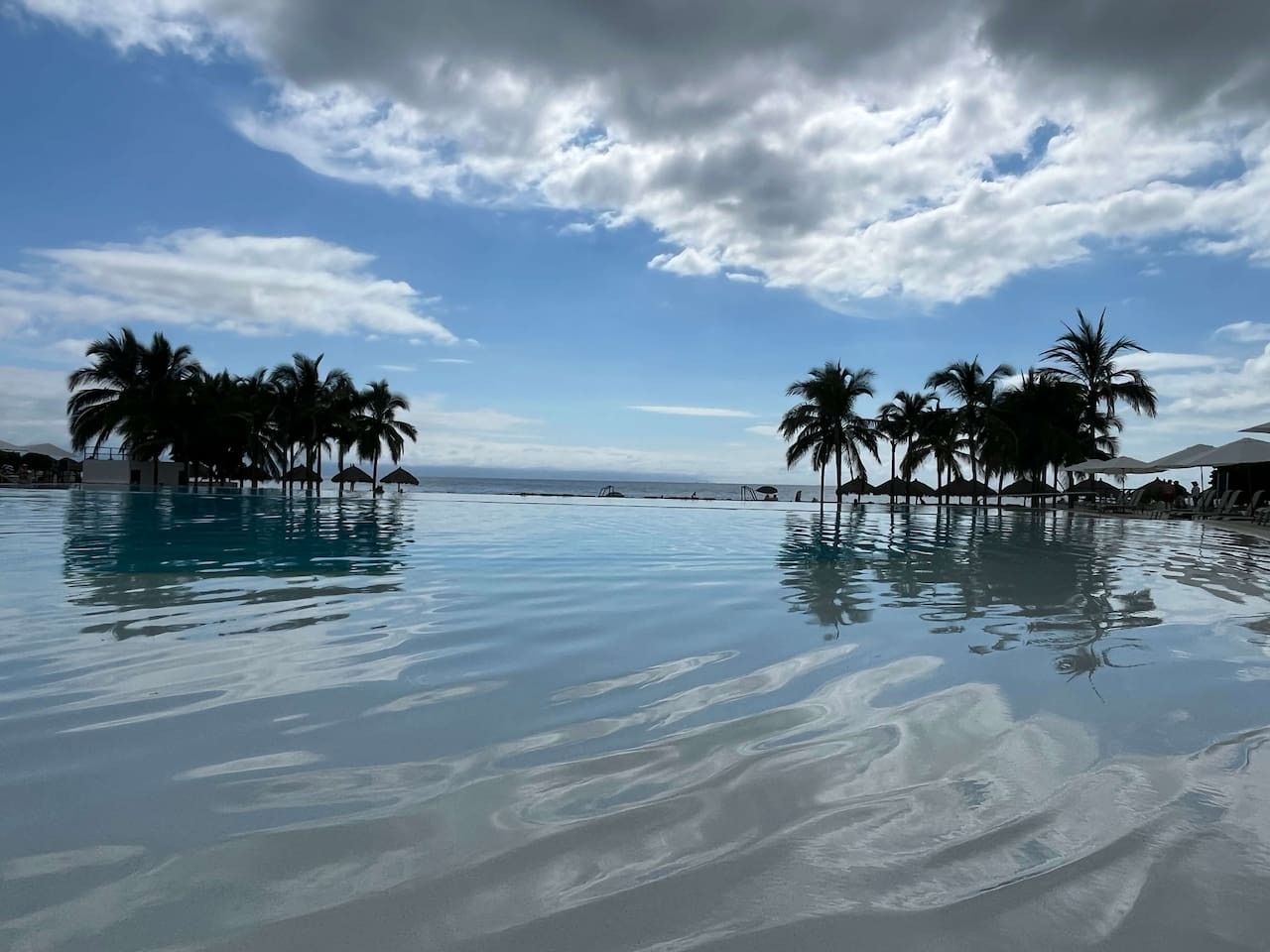 Cielo despejado con nubes, una piscina tranquila que refleja el entorno, y palmeras bordeando la orilla.