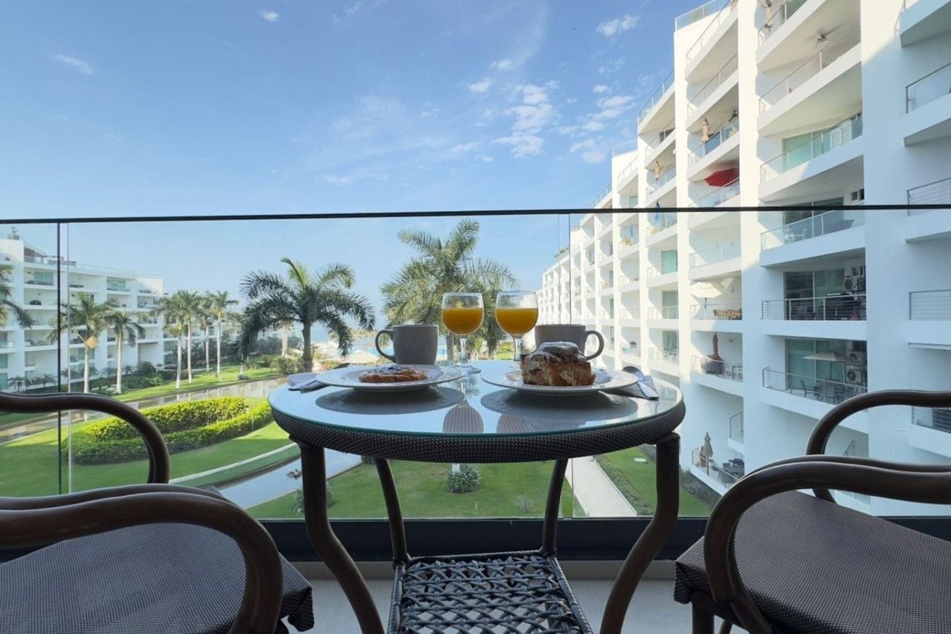 A balcony with a table set for breakfast, overlooking a modern building and a landscaped area with palm trees.