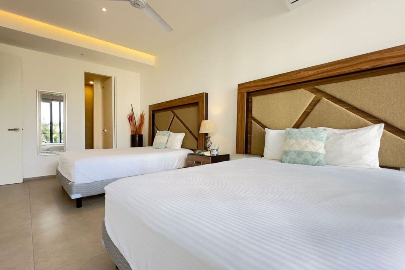 A hotel room with two beds, a ceiling fan, a window with a view, and a bedside table with a lamp.