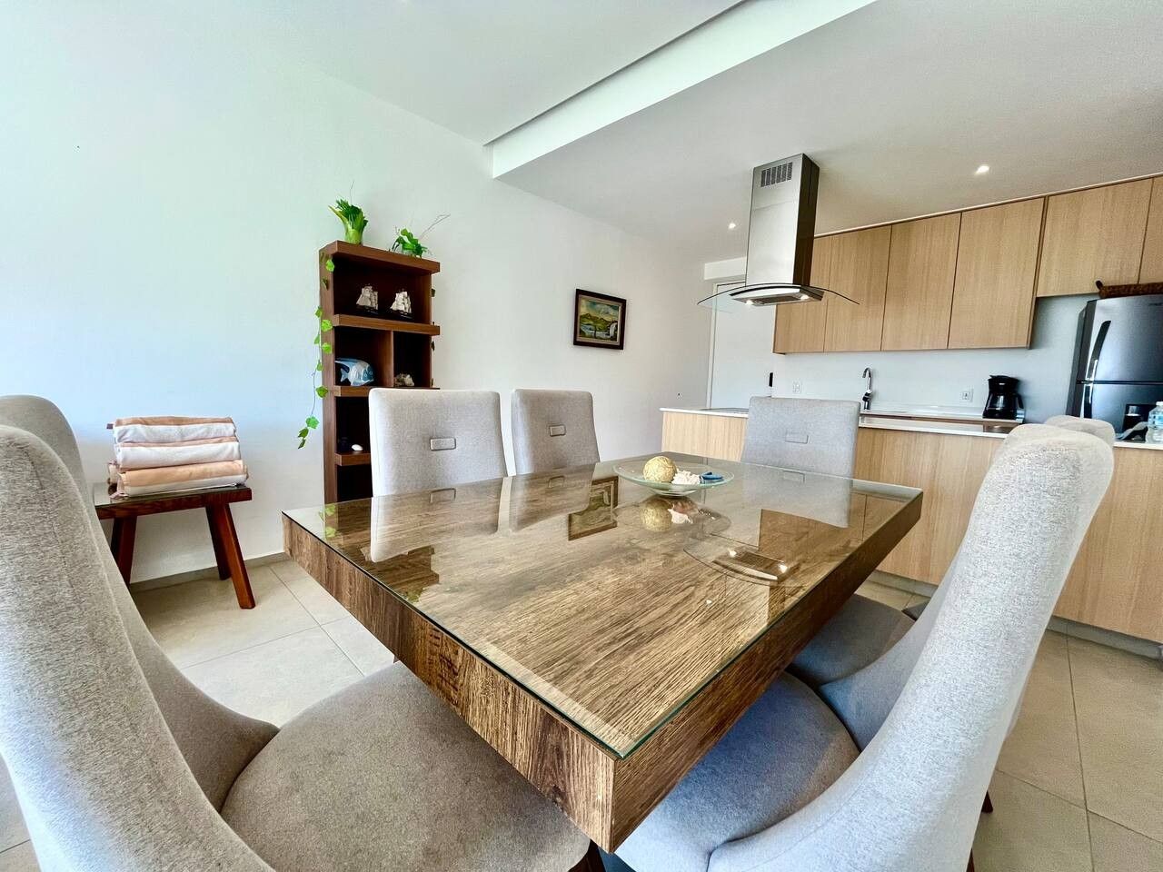 Un comedor moderno con una mesa de vidrio, sillas acolchadas, una mesita auxiliar con una planta y una cocina al fondo.