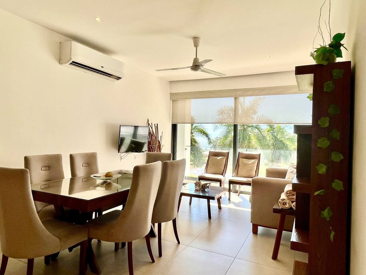 A modern dining area with a table and chairs, a ceiling fan, and a large window with a view of palm trees.