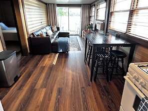 A compact living space with a sofa, a dining table with stools, a kitchen area with an oven, and a sliding door leading to an outdoor area.