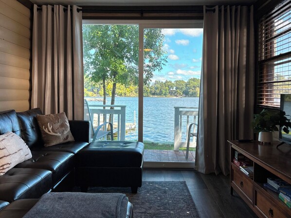 A living room with a black leather sofa, a wooden side table, and a view of a lake through a glass door.