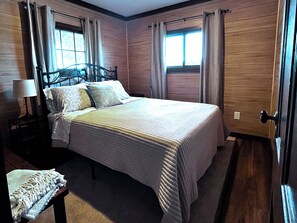 A bedroom with a bed, a nightstand, a lamp, and a window with curtains. - Charming Renovated Northwoods Cabin on Chetek Chain of Lakes (Chetek)