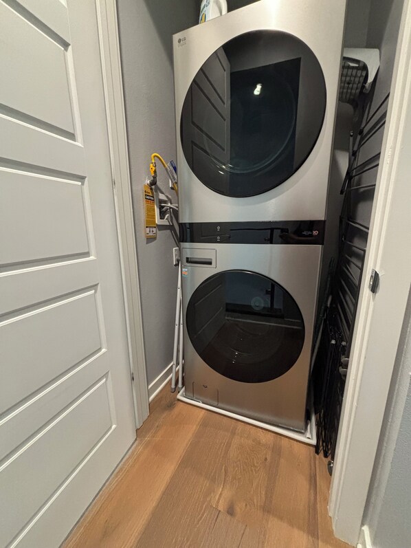 Une petite buanderie équipée d’un appareil combiné lave-linge et sèche-linge à deux portes, d’un panneau d’avertissement et d’une prise électrique bien visible.