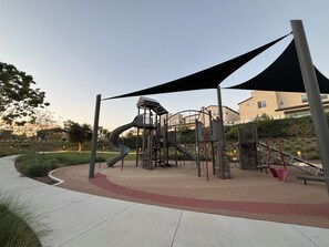 Une aire de jeux avec un toboggan et des structures d’escalade, entourée d’une allée pavée et de verdure.