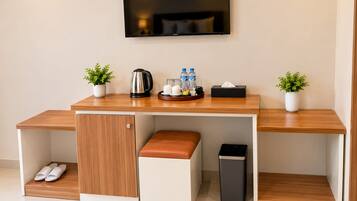 A modern hotel room with a wooden desk, a television mounted on the wall, a small seating area with a bench, and a trash bin.