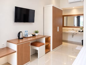 Quarto de hotel moderno com televisão de tela plana, escrivaninha de madeira com porta-copos, uma pequena planta e banheiro com pia e espelho.