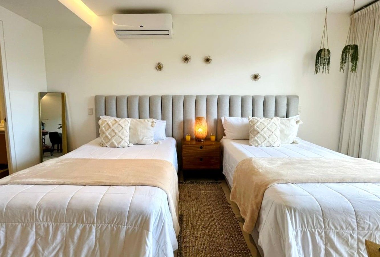 A hotel room with two beds, a nightstand with a lamp, and a wall-mounted air conditioner.