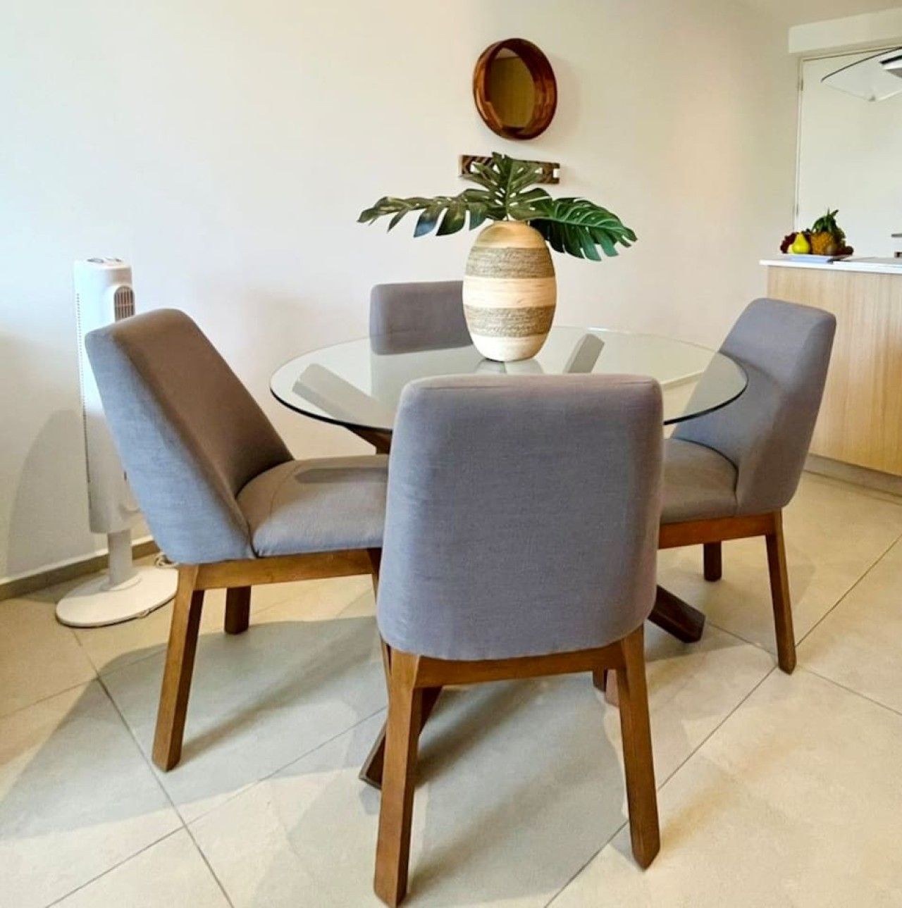 A dining area with a round glass table surrounded by four grey upholstered chairs with wooden legs.