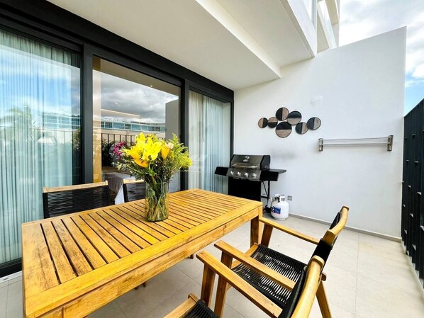 A balcony with a wooden table and chairs, a grill, and a vase with flowers. - Groundfloor condo w/grill beach & pools Aria Ocean (Nuevo Vallarta)