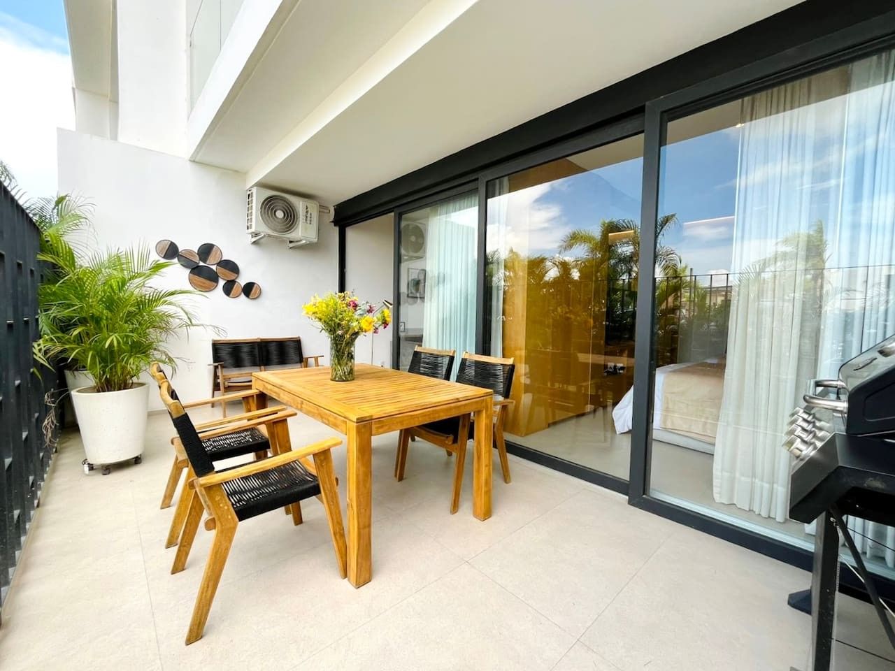 A modern outdoor dining area with a wooden table and chairs, a potted plant, and a view through a glass door.