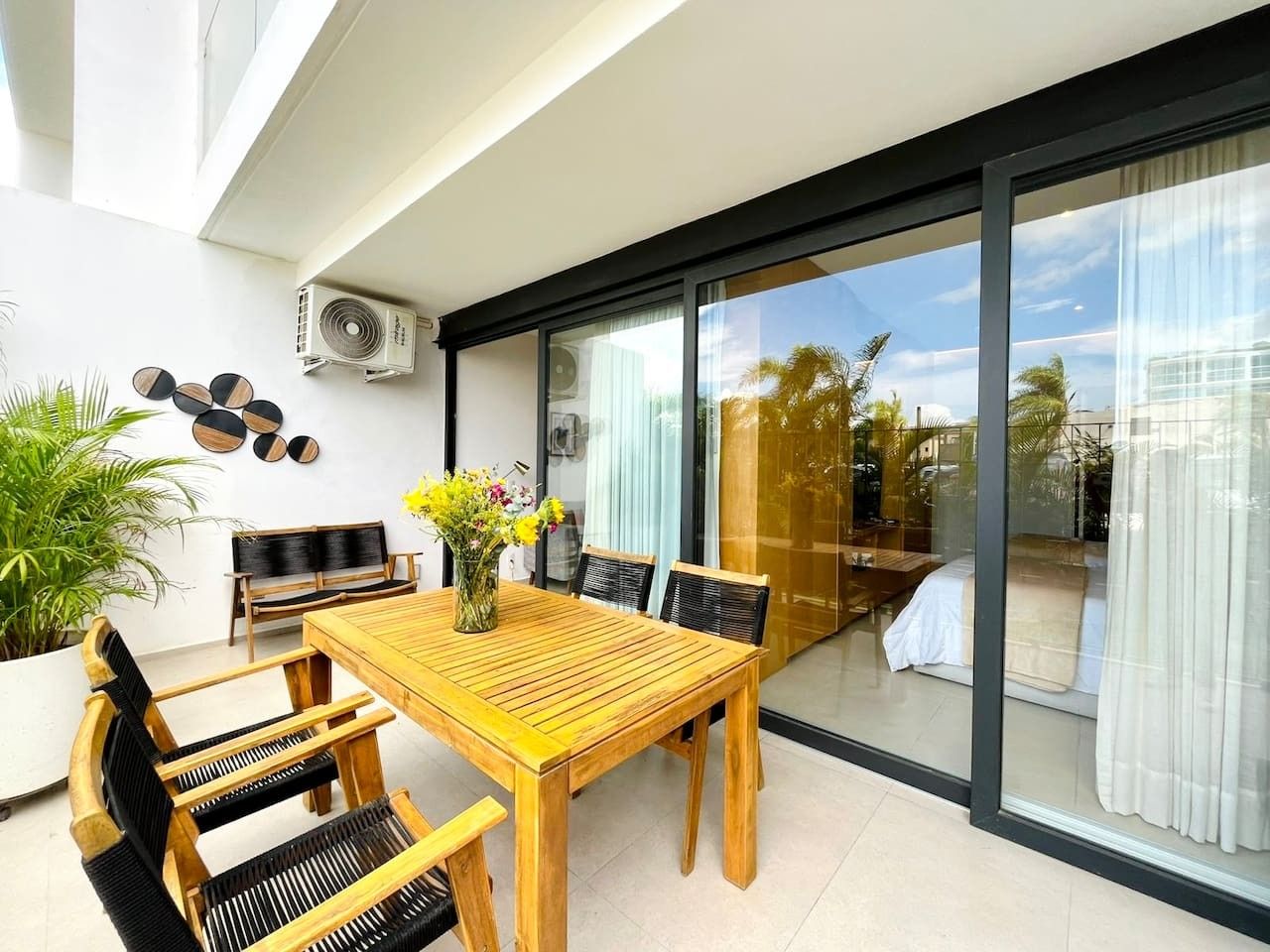 A modern dining area with a wooden table and chairs, a potted plant, and a view through a glass door to an outdoor space.