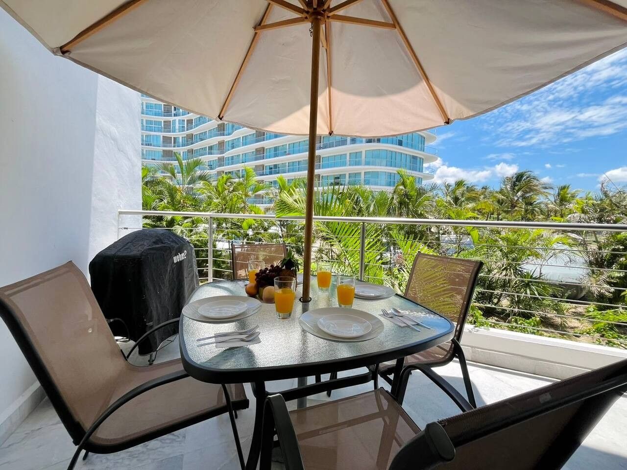Une table à plateau en verre, mise avec des assiettes, des couverts et des verres, placée sous un grand parasol sur un balcon d’où l’on peut admirer un immeuble d’hôtel et des palmiers.