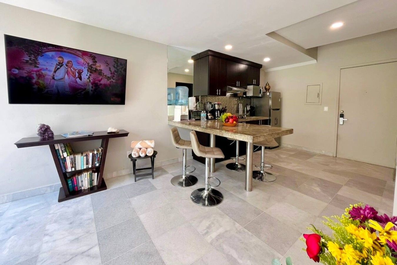 A modern kitchen with a dining area, a bookshelf, and a painting on the wall.