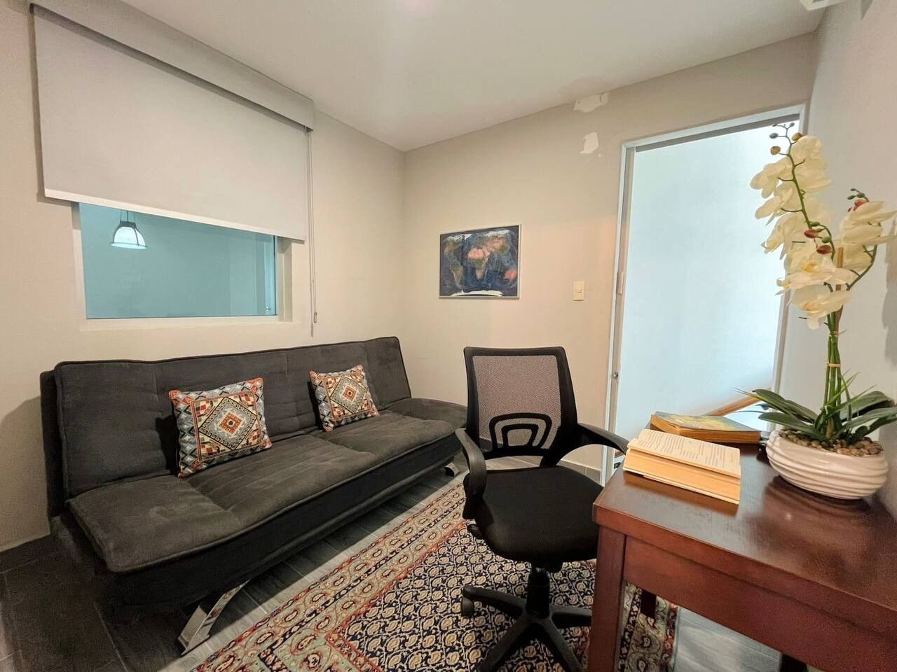 Un salon avec un canapé, un fauteuil, une petite table sur laquelle est posée une plante, et une fenêtre avec des stores.