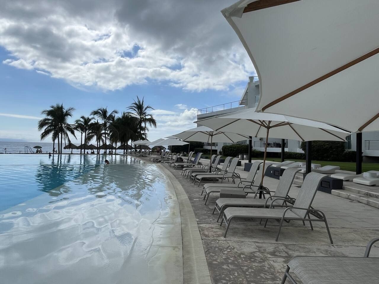 Área da piscina com espreguiçadeiras e guarda-sóis, palmeiras e céu claro.