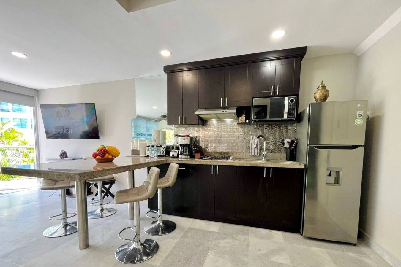 A modern kitchen with dark cabinetry, a stainless steel refrigerator, and a flat-screen TV mounted on the wall.