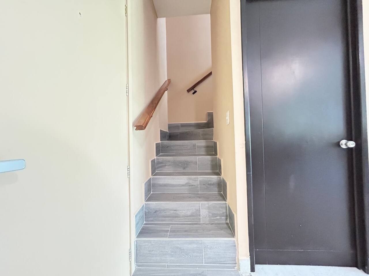 Escada que leva a uma porta escura, com uma parede clara de um lado e um corrimão de madeira.
