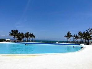Ein klares, blaues Schwimmbecken mit Palmen und Liegestühlen, umgeben von einem weißen Sandstrand.