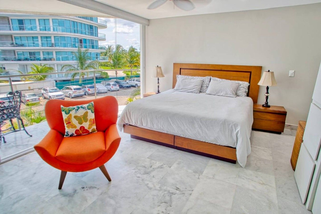 A hotel room with a bed, a chair, a nightstand, and a view of a building and parking area.