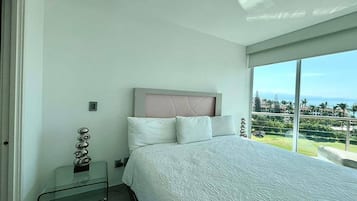 A bedroom with a large bed, a glass bedside table, a headboard, and a view of a green landscape through large windows.