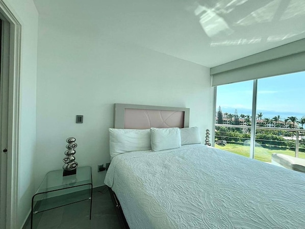 A bedroom with a large bed, a glass bedside table, a headboard, and a view of a green landscape through large windows. - 520 sea view apartment in Nuevo Vallarta (Nuevo Vallarta)