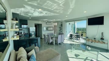 A modern living room with a glass coffee table, a sofa with cushions, a dining area with a glass table, and a kitchen with a built-in oven.