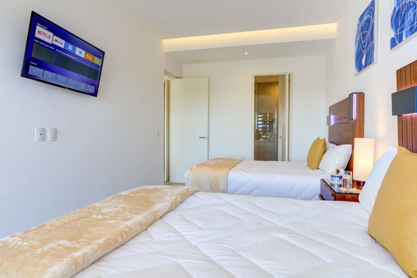 A hotel room with two beds, a TV mounted on the wall, and a bedside table with a lamp.