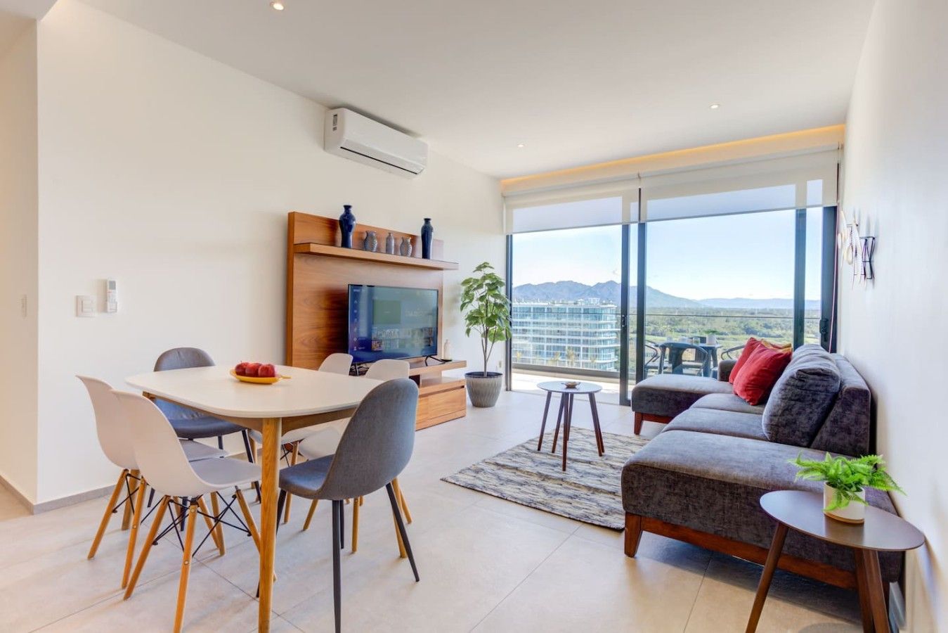 A modern living room with a dining area, a sofa, and a TV. There is a large window with a view of the mountains.