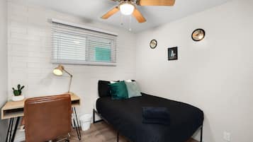Un dormitorio compacto con cama, escritorio con silla, ventilador de techo, ventana con persianas y una pequeña planta.