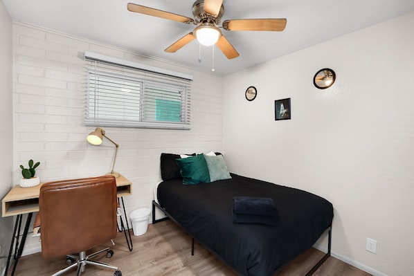 Un dormitorio compacto con cama, escritorio con silla, ventilador de techo, ventana con persianas y una pequeña planta.