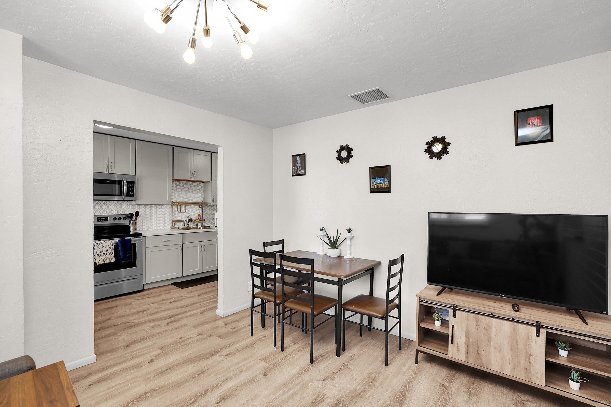 Una cocina moderna con armarios blancos y electrodomésticos de acero inoxidable, un comedor con mesa y sillas de madera, y un televisor de pantalla plana grande sobre un mueble de madera.