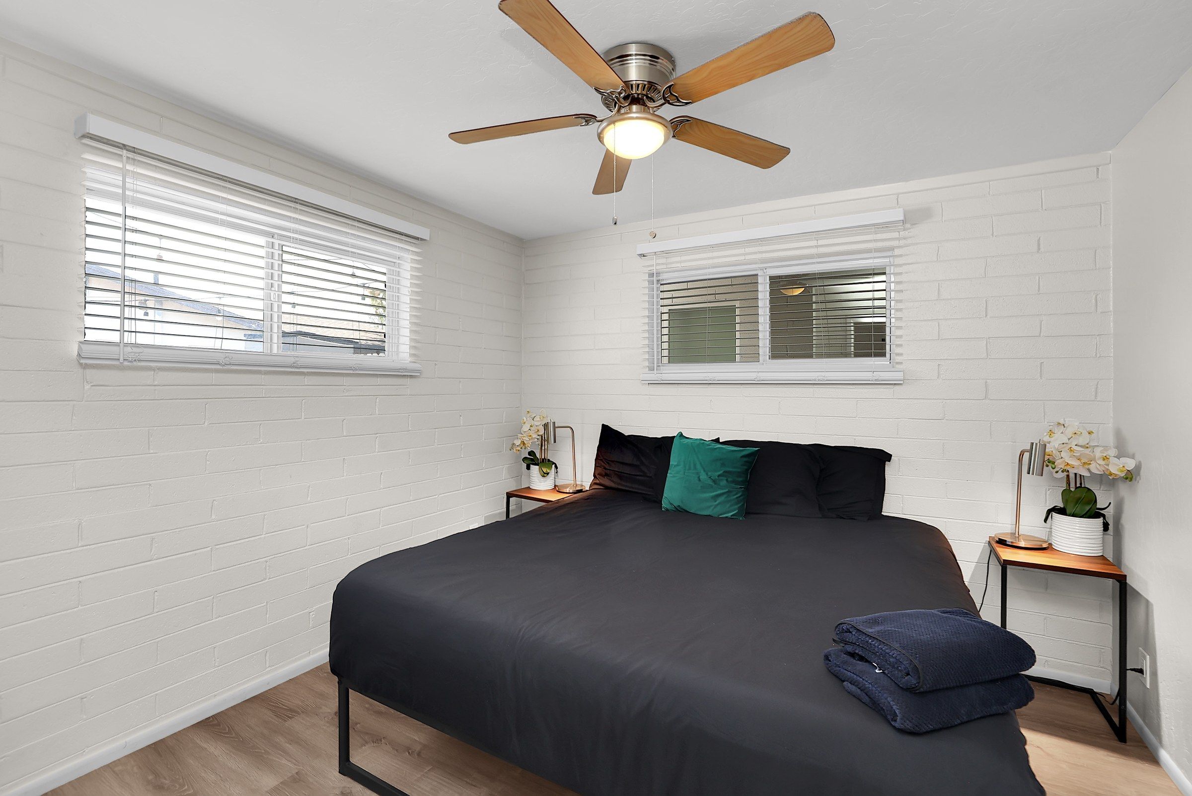 Un dormitorio con una cama, dos mesitas de noche, un ventilador de techo y ventanas con persianas.