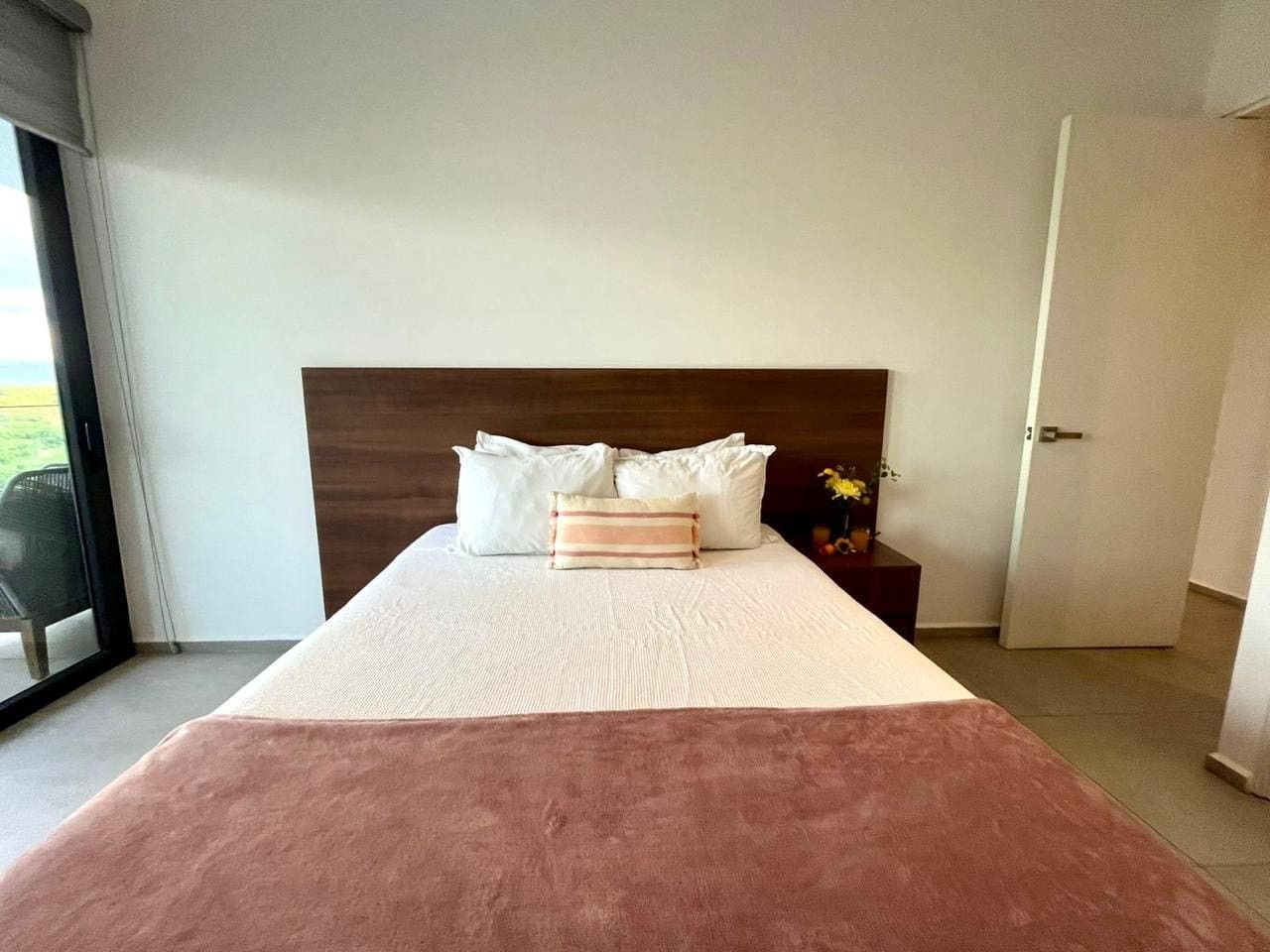 A hotel room with a large bed, a wooden headboard, a nightstand with a vase of flowers, and a sliding glass door leading to a balcony.