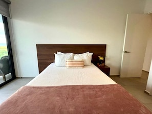 Quarto de hotel com uma cama grande, cabeceira de madeira, mesa de cabeceira com um vaso de flores e uma porta de vidro deslizante que dá para uma varanda.