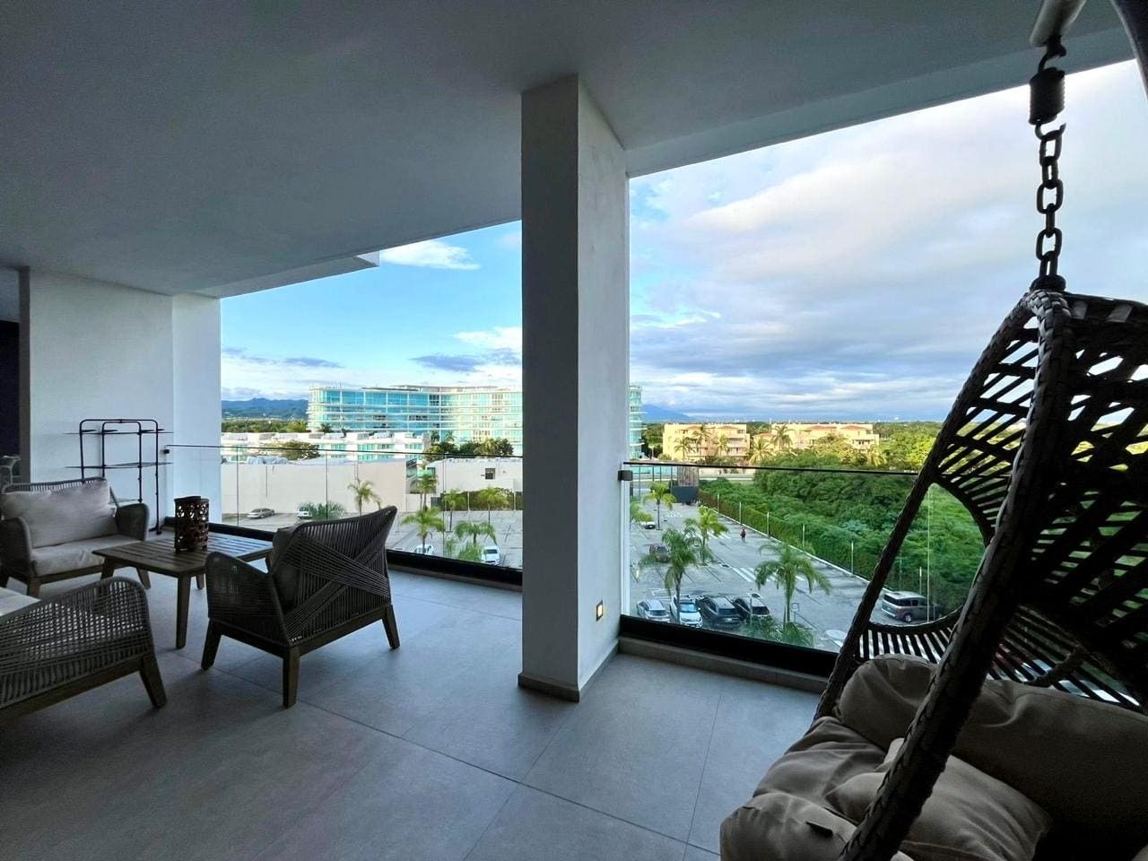 Un balcon aménagé avec un canapé, des chaises et un fauteuil suspendu. On y aperçoit une vue sur le paysage urbain et la verdure.