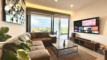A modern living room with a large flat-screen TV, a sofa, a coffee table, and a large window with a view.
