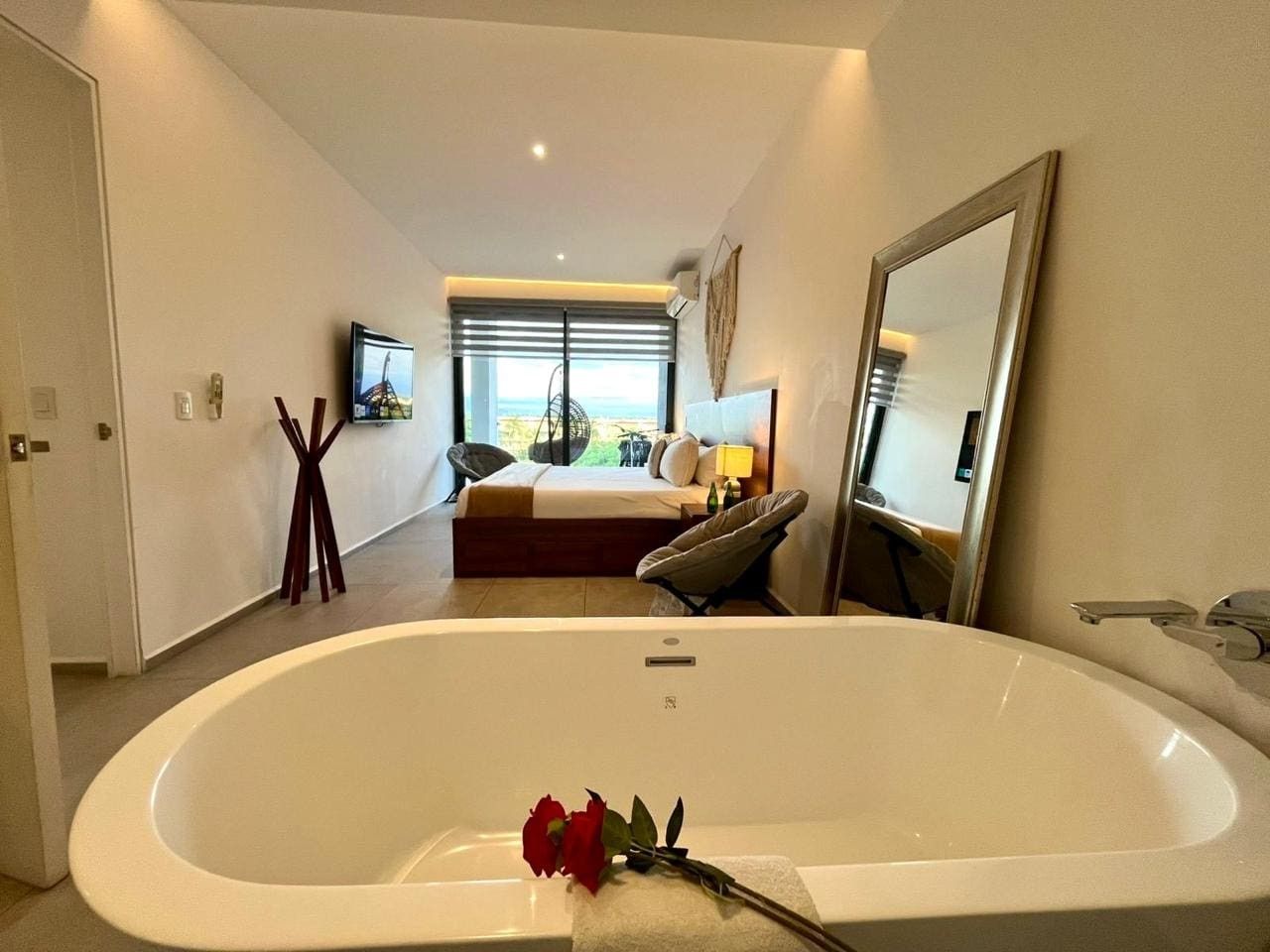 A modern bathroom with a large bathtub, a mirror, and a bed in the background.