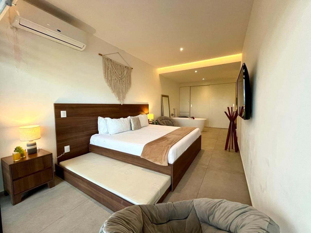 Un dormitorio moderno con una cama grande, mesitas de noche, un televisor montado en la pared y un baño al fondo.