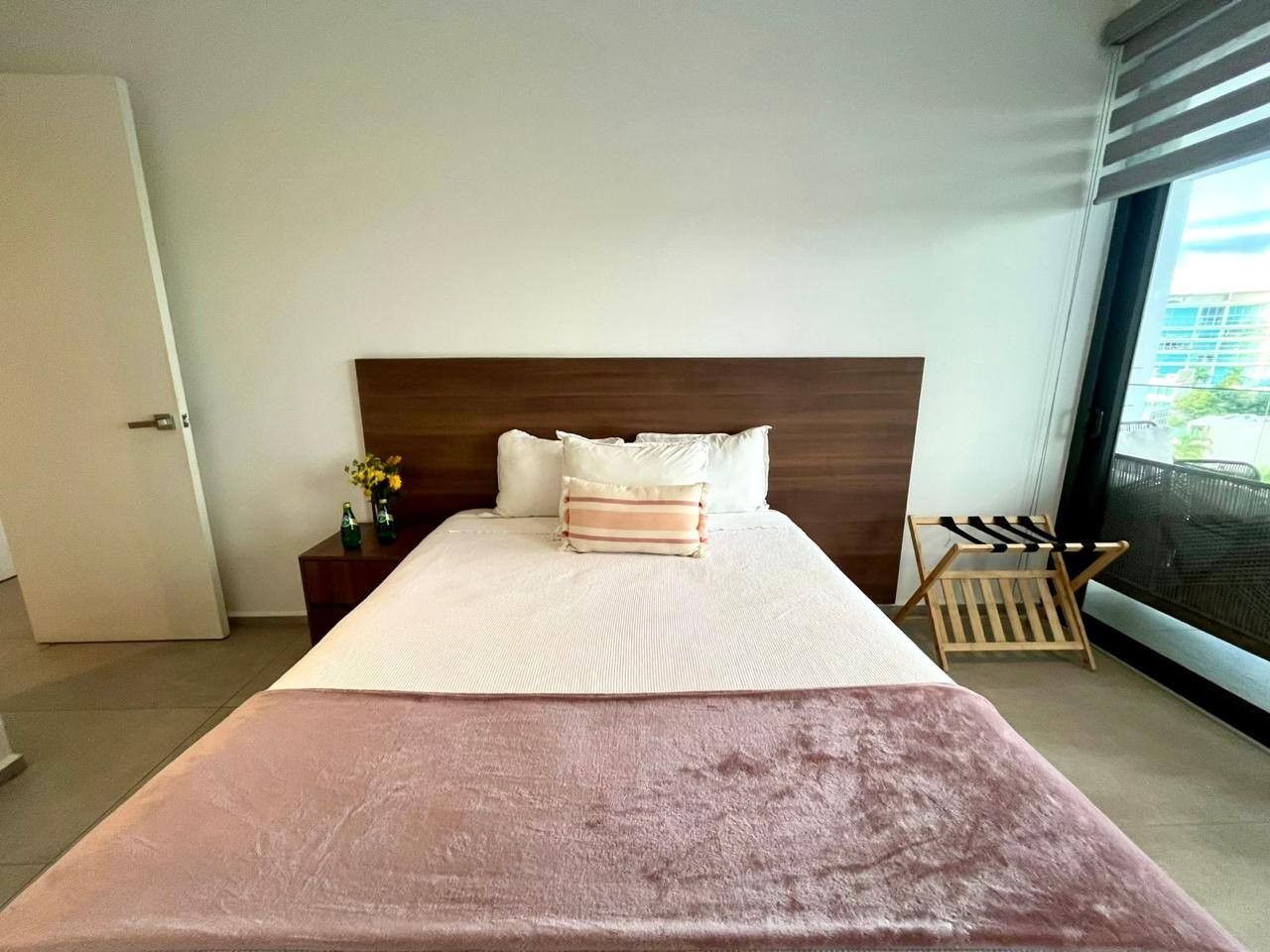 A hotel room with a large bed, a wooden headboard, a nightstand with a vase of flowers, and a window with a view of buildings.
