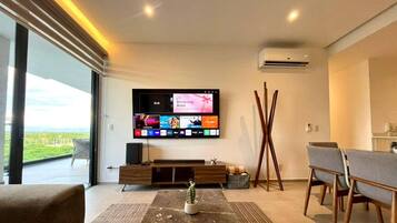 Uma sala de estar moderna com uma TV de tela plana, um cacto como peça central, e uma área de jantar com cadeiras e uma mesa.