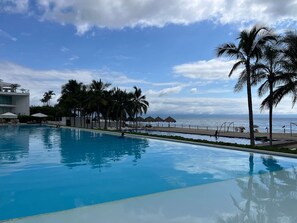 Una piscina con vista al océano, palmeras y una zona de playa con sombrillas.
