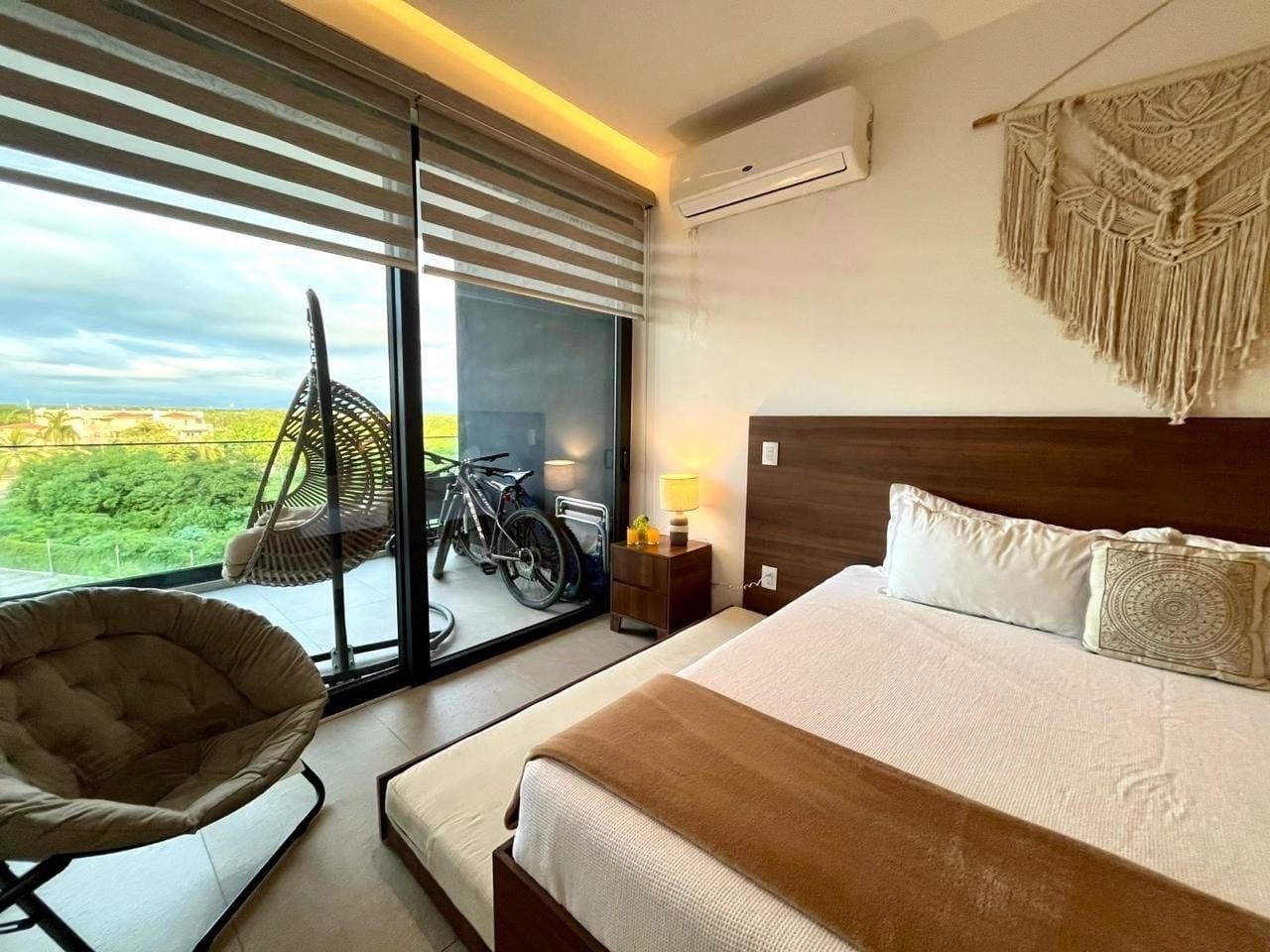 A modern hotel room with a large bed, a chair, a view of a bicycle, and a macrame wall hanging.