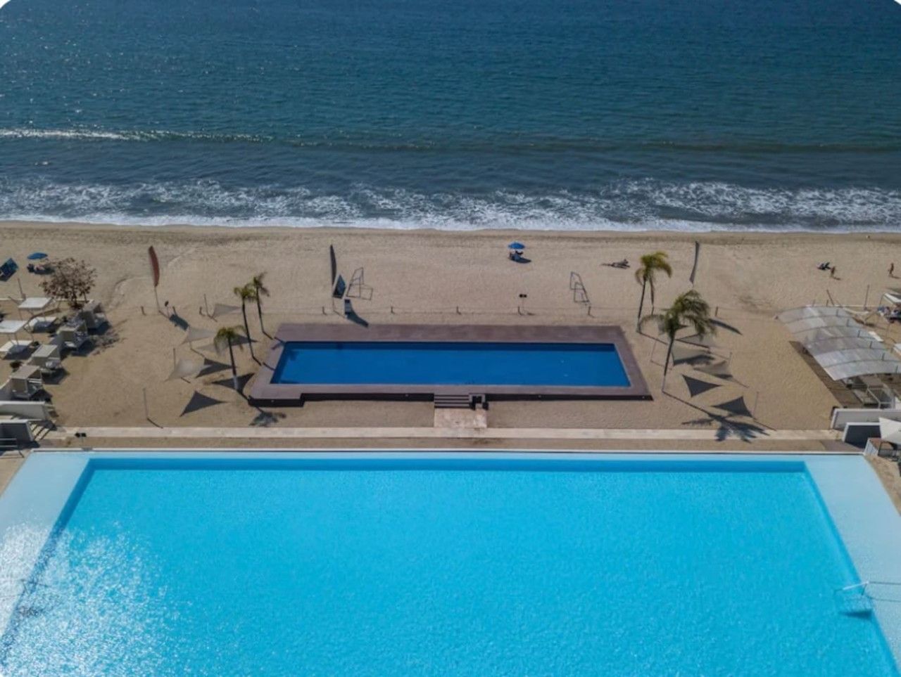 A swimming pool adjacent to a beach with lounge chairs and umbrellas.