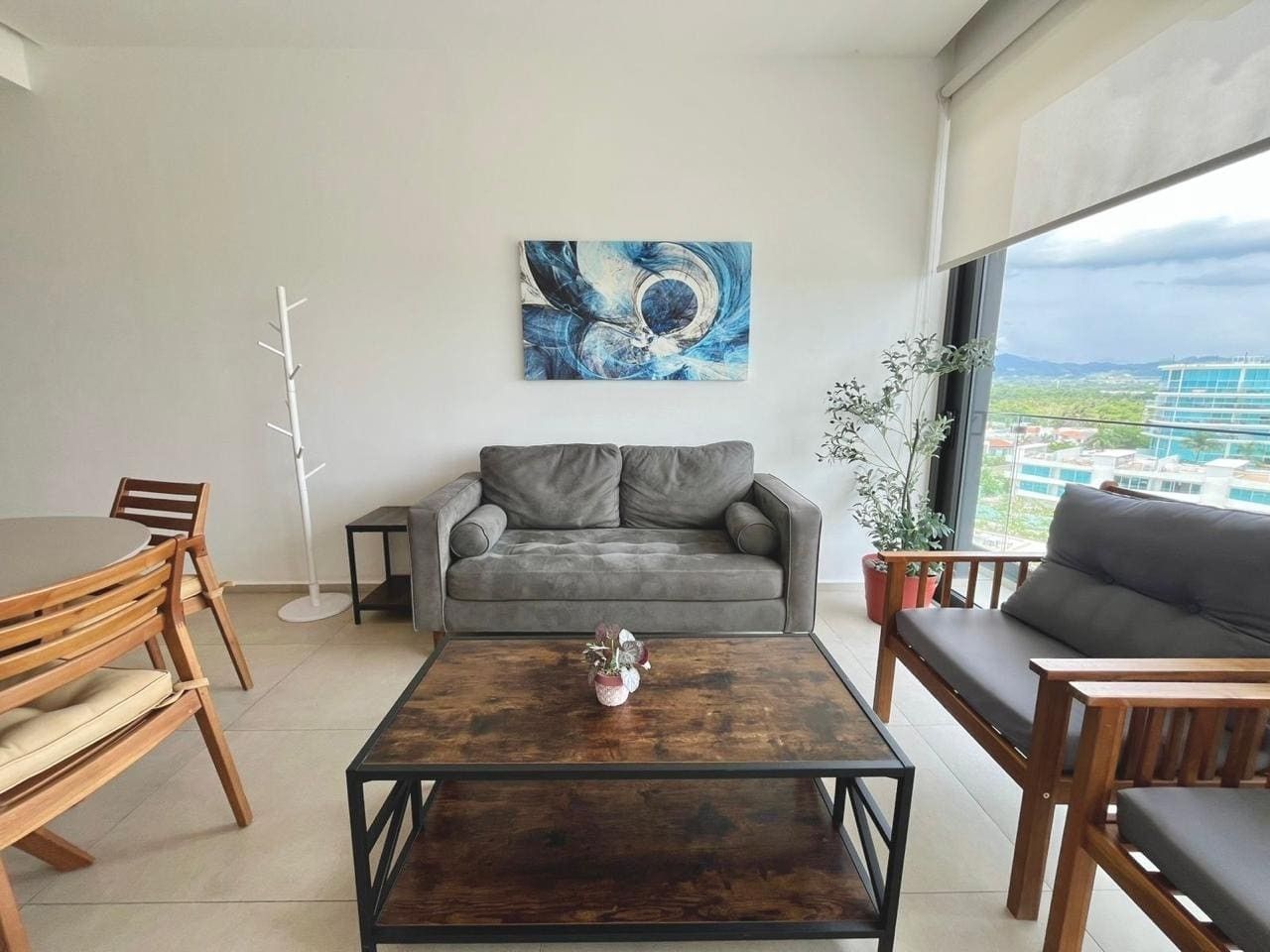 Een moderne woonkamer met een grijze bank, houten stoelen, een salontafel en een groot raam met uitzicht.