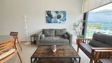 Een moderne woonkamer met een grijze bank, houten stoelen, een salontafel en een groot raam met uitzicht.